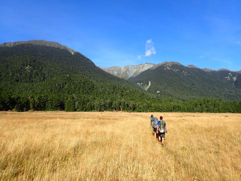 3 hikers crossing grassy valley flats in the sunshine with mountains beyond