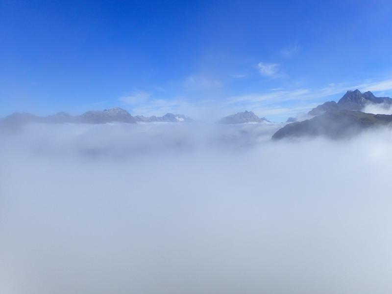 blue sky above and clouds below with a few mountain peaks rising above the clouds