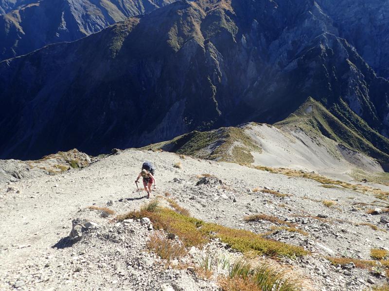 a hiker climbing up pale gravel and rocks, further below is a mountain saddle