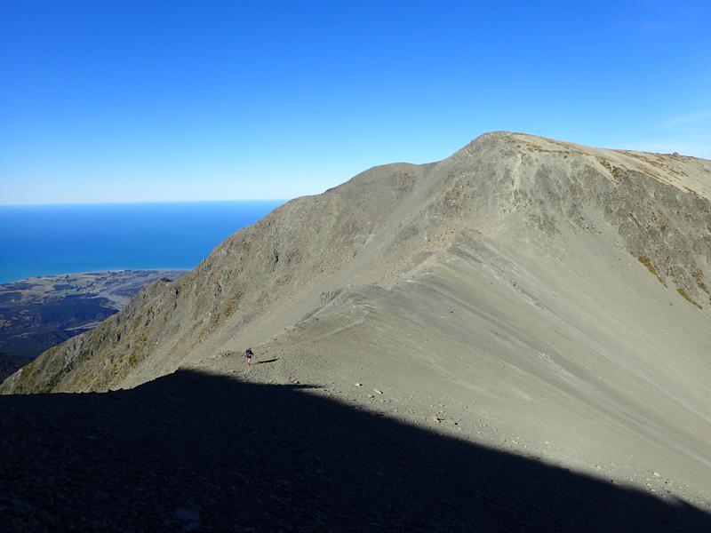 a hiker crossing a large scree covered ridge between two peaks, the sea can be seen in the background