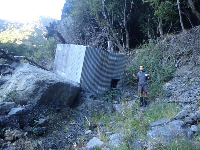 a corrugated tin hut sitting on rocks upside down, with a hiker holding an unbroken mug and glass