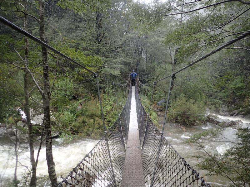 a narrow swing bridge over a river with trees beyond