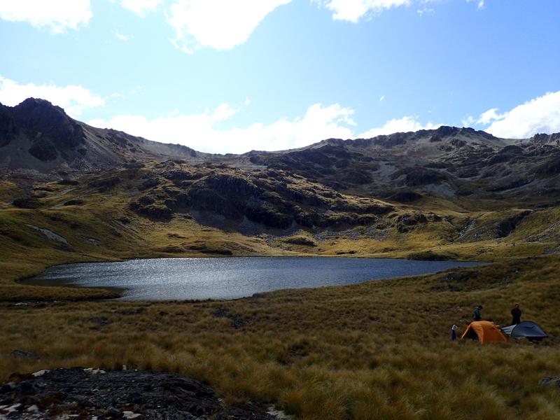 a lake with 2 tents and hikers and mountains behind