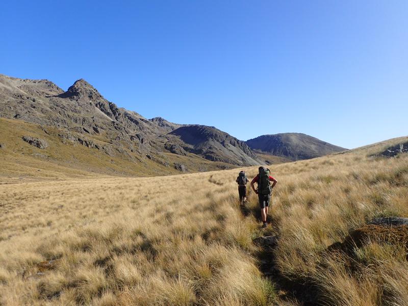 2 hikers walking along a grassy valley with mountains beyond