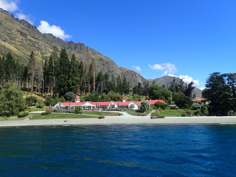 Grand white building with red roof and smart gardens, trees then mountains behind
