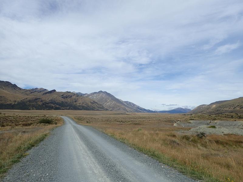 Straight gravel road through tussock with mountains behind
